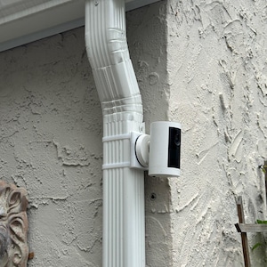 May include: A white cylindrical security camera mounted to a white vertical metal downspout using a custom bracket. The camera features a black circular lens area on its front face and is positioned against a textured light-colored exterior wall.
