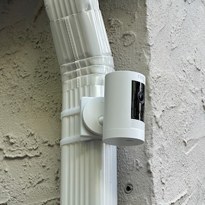 May include: A white cylindrical security camera with the brand name Ring printed on the top edge. The device features a black circular lens area and a perforated speaker section, mounted to a white vertical downspout using a matching bracket.