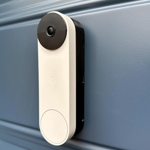May include: A white and black smart doorbell with a camera lens and the Google logo. The doorbell is mounted on a blue surface.