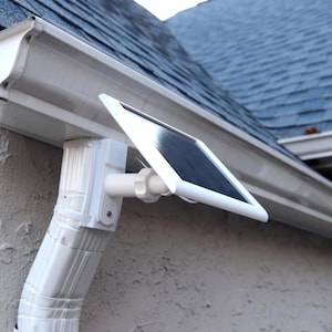 May include: A white solar-powered gutter light is mounted on a white gutter system. The light has a rectangular solar panel and is attached to the gutter with a white bracket. The background includes a blue shingled roof and a stucco wall.