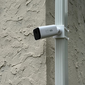 May include: A white Eufy 2K Security camera mounted to a vertical white downspout. The camera has a rectangular body with a black front face and is attached to the downspout using a white plastic bracket secured with zip ties.