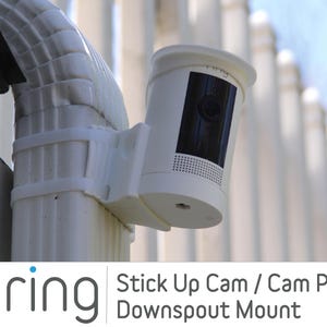 May include: White Ring Stick Up Cam or Cam Pro mounted on a white downspout with a white bracket. The camera is pointed towards the right. The text "ring" is visible on the camera.