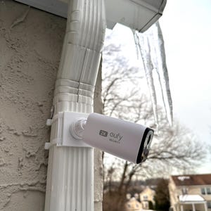 May include: A white security camera labeled "2K eufy SECURITY" is mounted on a white gutter. The camera is cylindrical with a black lens and is attached to a white bracket. The background includes a snowy landscape and a house.