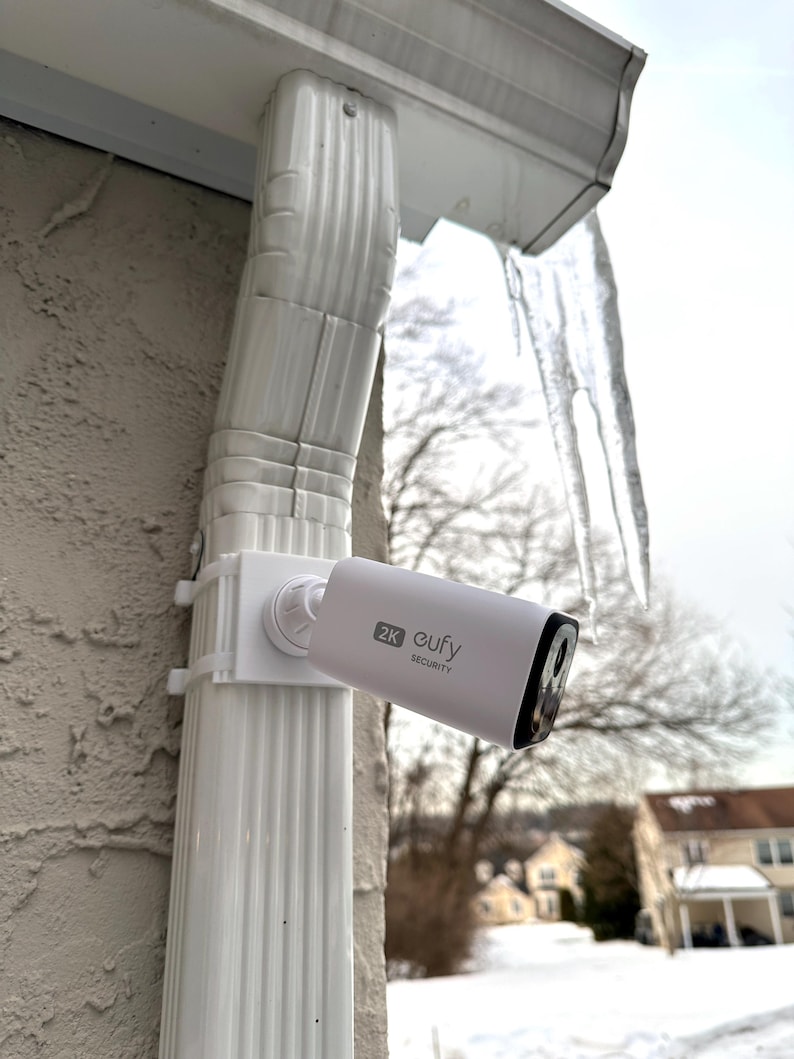 May include: A white security camera labeled "2K eufy SECURITY" is mounted on a white gutter. The camera is cylindrical with a black lens and is attached to a white bracket. The background includes a snowy landscape and a house.