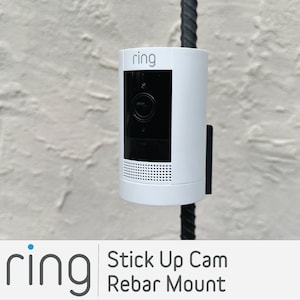 May include: A white Ring Stick Up Cam mounted on a black rebar with the text "ring" on the front of the camera.