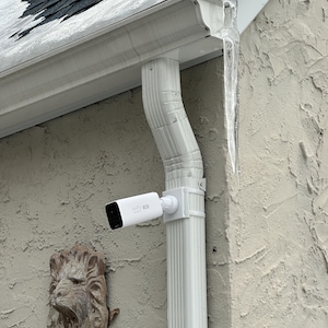 May include: A white security camera is mounted on a white downspout. The camera is cylindrical with a black front. The downspout is attached to a textured, light-colored wall. A decorative lion's head is mounted on the wall.