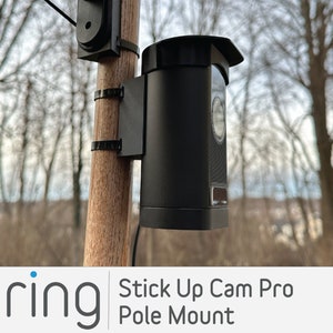 May include: A black Ring Stick Up Cam Pro mounted on a wooden pole with a black pole mount. The camera is pointed towards the right. The background is blurred and shows trees.