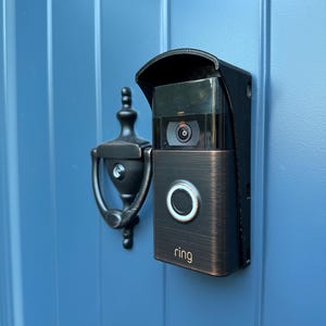 May include: A bronze-colored Ring doorbell with a black visor and a white button. The doorbell is mounted on a blue door.
