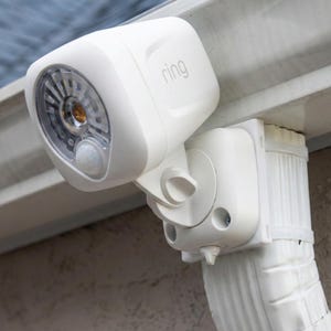 May include: A white Ring security floodlight with a motion sensor. The light is mounted on a white bracket and attached to a white downspout. The Ring logo is visible on the top of the light.