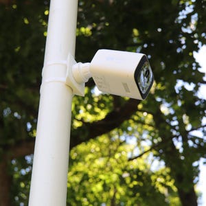 May include: A white security camera mounted on a vertical white pole. The camera has a rectangular white housing with a black front face containing the lens. The mounting bracket connects the camera to the pole with a circular adjustment joint.