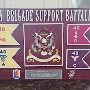 Może przedstawiać: Burgundowa drewniana tablica z białym napisem "40TH BRIGADE SUPPORT BATTALION". Zawiera emblematy wojskowe i nazwisko "CSM IRMA ORTIZ". Tablica ma złote, białe i inne kolorowe akcenty.