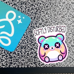 Może przedstawiać: Naklejka z kresk&oacute;wkowym niedźwiedziem w kolorze r&oacute;żowym, niebieskim i zielonym. Niedźwiedź ma smutny wyraz twarzy z X zamiast oczu i łzami, kt&oacute;re kapia. Tekst "Softly Disturbed" znajduje się nad niedźwiedziem.
