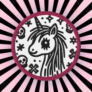Op de afbeelding: Een witte geborduurde patch met een zwarte lijntekening van een eenhoorn met een enkele hoorn en een schedel en gekruiste beenderen ontwerp. De patch heeft een roze rand en is geplaatst tegen een zwart-roze gestreepte achtergrond.