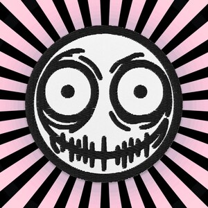Peut inclure: Un patch brodé noir et blanc représentant un visage de dessin animé avec de grands yeux et un sourire narquois. Le patch est placé sur un fond rayé rose et noir.