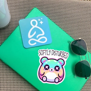 Może przedstawiać: Zielony notes z naklejką z kresk&oacute;wkowym bohaterem z tekstem "Softly Disturbed" i niebieską naklejką ze stylizowaną postacią siedzącą w pozycji lotosu z tekstem "Softly Disturbed".