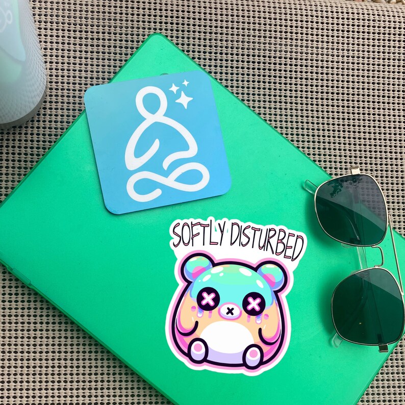 Może przedstawiać: Zielony notes z naklejką z kresk&oacute;wkowym bohaterem z tekstem "Softly Disturbed" i niebieską naklejką ze stylizowaną postacią siedzącą w pozycji lotosu z tekstem "Softly Disturbed".
