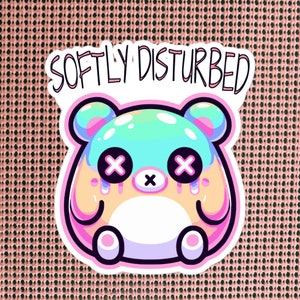 Może przedstawiać: Naklejka przedstawiająca kresk&oacute;wkowego niedźwiedzia z ciałem w pastelowych kolorach niebieskim i zielonym, r&oacute;żowymi uszami i białym futrem. Niedźwiedź ma duże oczy z czarnymi X i r&oacute;żowy nos. Naklejka ma r&oacute;żową ramkę i napis "SOFTLY DISTURBED" w kolorze czarnym.