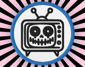 May include: A black and white embroidered patch of a television with a skull face. The patch is circular and has a blue border. The background is a pink and black striped pattern.