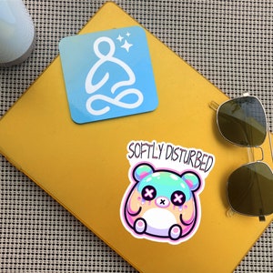 Może przedstawiać: Ż&oacute;łte tło z naklejką przedstawiającą pastelowo-niebieską i r&oacute;żową postać z kresk&oacute;wki z tekstem "Softly Disturbed" nad nią. Jest niebieska naklejka z białym konturem osoby siedzącej w pozycji medytacyjnej z gwiazdami nad nią.