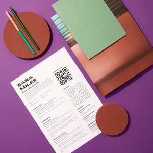 Op de afbeelding: Een cv met de naam "SARA MILES" en de titel "SOCIAL MEDIA MANAGER" bovenaan. Het cv ligt op een paarse ondergrond met twee bruine onderzetters en twee potloden. Het cv bevat een QR-code, vaardigheden, ervaring, opleiding en onderscheidingen.
