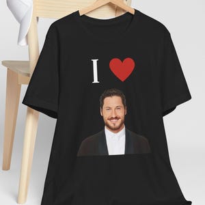 May include: Black t-shirt with the text "I ♥" and a red heart graphic. Below the text is a smiling man wearing a black suit and white shirt. The shirt is hanging on a wooden hanger.