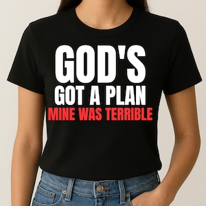 Peut inclure: Un t-shirt noir à manches courtes avec le texte blanc "GOD'S GOT A PLAN" sur les deux premières lignes et le texte rouge "MINE WAS TERRIBLE" sur la ligne du bas. Le t-shirt est associé à un jean bleu avec un bouton en métal visible.