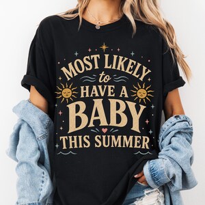 Peut inclure: T-shirt noir avec l'inscription "MOST LIKELY to HAVE A BABY THIS SUMMER" dans une police fantaisiste, ornée de soleils et d'étoiles. La personne porte une veste en jean.