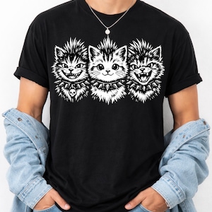 May include: Black t-shirt featuring a graphic of three stylized cat faces with spiky collars. The cats have mohawk-style fur and varying expressions, with one wearing a skull pendant. The design is in black and white.