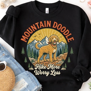 Peut inclure: Sweat-shirt noir avec un motif de chien golden doodle dans un paysage de montagne. Le texte "Mountain Doodle" et "Hike More Worry Less" est inclus. Le design comprend des montagnes, des arbres et un soleil.