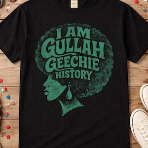 May include: Black t-shirt with a green graphic design. The design features the text "I AM GULLAH GEECHIE HISTORY" in a retro font, along with a silhouette of a woman's profile with an afro and a teardrop earring. The shirt is on a wooden surface.