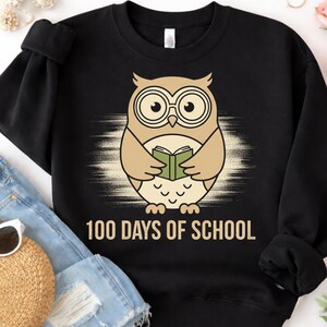 Peut inclure: Sweat-shirt noir avec un dessin de hibou de dessin animé. Le hibou, de couleur beige et marron, porte des lunettes et tient un livre. Le texte "100 DAYS OF SCHOOL" est imprimé en blanc.