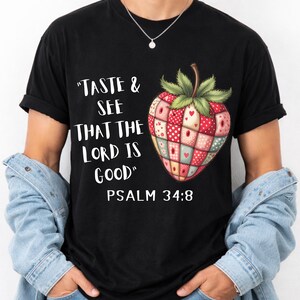 Peut inclure: T-shirt noir avec une fraise colorée en patchwork. Le texte "TASTE & SEE THAT THE LORD IS GOOD" et "PSALM 34:8" est imprimé. La fraise est composée de carrés rouges, roses et verts.