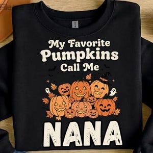 Peut inclure: Un sweat-shirt noir à manches longues avec une impression graphique portant le texte "My Favorite Pumpkins Call Me NANA". Le motif comprend plusieurs citrouilles orange sculptées, des feuilles d'automne, des chauves-souris et deux fantômes blancs.
