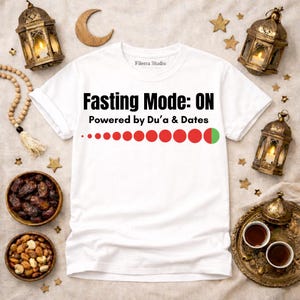Peut inclure: T-shirt blanc avec le texte "Fasting Mode: ON" en noir et "Powered by Du'a & Dates" en dessous. Une série de points rouges et un demi-cercle vert sont en dessous. Le t-shirt est entouré de dattes, de noix, de lanternes et d'autres décorations.