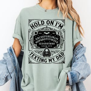 May include: A light green t-shirt with a black Ouija board design. The design includes the words "HOLD ON I'M TEXTING MY DAD" and a decorative frame with letters, numbers, and a planchette. The shirt has rolled-up sleeves.