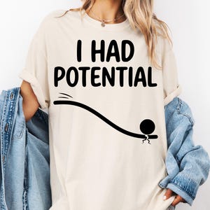 Pode incluir: Camiseta creme com a frase "I HAD POTENTIAL" em letras pretas em negrito. Abaixo, um gráfico preto de uma bola rolando por uma linha curva. A camiseta tem um corte folgado e casual.