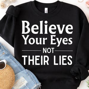 Puede incluir: Sudadera negra con el texto blanco "Believe Your Eyes NOT THEIR LIES". La sudadera se muestra con unos pantalones cortos de mezclilla azules, un sombrero de paja y gafas de sol.