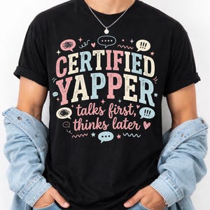 May include: Black t-shirt with the text "CERTIFIED YAPPER talks first, thinks later" in pastel colors. The design includes speech bubbles, stars, and wavy lines. The shirt is worn with a denim jacket.