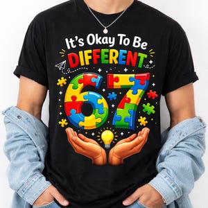 Peut inclure: T-shirt noir avec l'inscription colorée "It's Okay To Be DIFFERENT". Le chiffre "67" est composé de pièces de puzzle, avec une ampoule tenue dans des mains ouvertes. Un message d'acceptation et d'individualité.
