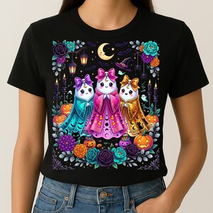 Peut inclure: Un t-shirt noir avec un motif coloré représentant trois fantômes portant des nœuds et des capes bleu sarcelle, rose et dorées. Le design inclut des citrouilles, des fleurs, des lanternes, un croissant de lune, un chapeau de sorcière et des chats noirs.