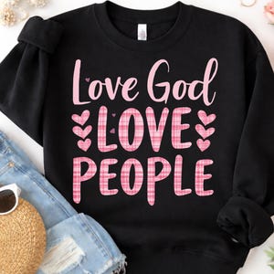 Peut inclure: Sweat-shirt noir avec l'inscription rose "Love God LOVE PEOPLE". Les mots sont écrits en script et en carreaux, avec des cœurs roses. Le sweat-shirt est associé à un short en jean et un chapeau de paille.