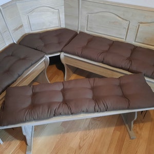 May include: A light-colored wooden corner bench with four brown cushions. The bench has a classic design with decorative panels and curved legs. The cushions are rectangular and tufted, providing comfortable seating.