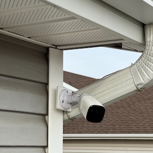 May include: A white and black security camera mounted on a white bracket, attached to a house's exterior. The camera is positioned under the roof's overhang, near a gutter and downspout. The house has gray siding.