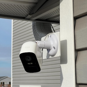 May include: A white security camera mounted on a gray exterior wall. The camera has a black lens and is attached to a white bracket. The house has gray siding and a white trim. The camera is installed under an overhang.