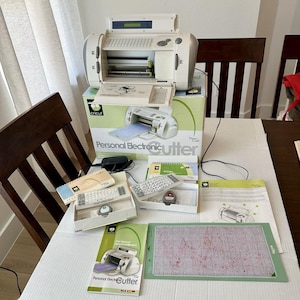May include: A beige Cricut Personal Electronic Cutter machine sits atop its original box. Included are two white keypads, a power adapter, two spiral-bound instruction booklets, a user manual, and a green rectangular cutting mat with grid lines.