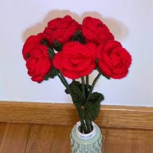 Puede incluir: Un ramo de rosas rojas de ganchillo en un jarrón azul claro con un patrón texturizado. Las rosas tienen tallos y hojas verdes. El jarrón es cilíndrico y está sobre un suelo de madera.