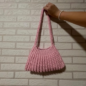 Puede incluir: Un bolso rosa de ganchillo hecho a mano con una forma trapezoidal estructurada. El bolso presenta una correa larga trenzada y una abertura superior redondeada. El bolso está hecho de un hilo suave y texturizado.