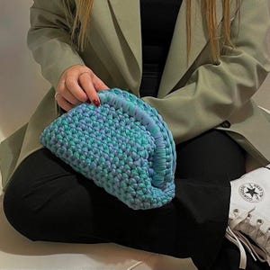 May include: A blue and green crocheted handbag with a half-circle shape. The bag is being held by a person wearing a green blazer and black pants.