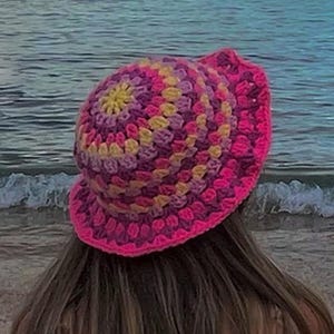 Puede incluir: Un sombrero de cubo de crochet rosa, amarillo y morado. El sombrero está hecho de un hilo suave y texturizado y tiene un ala ancha.