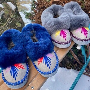 May include: Two pairs of cozy, handcrafted slippers. One pair features vibrant blue fur trim, while the other has gray fur. Both have embroidered designs in red, blue, and yellow on a light gray base, with stitched edges.